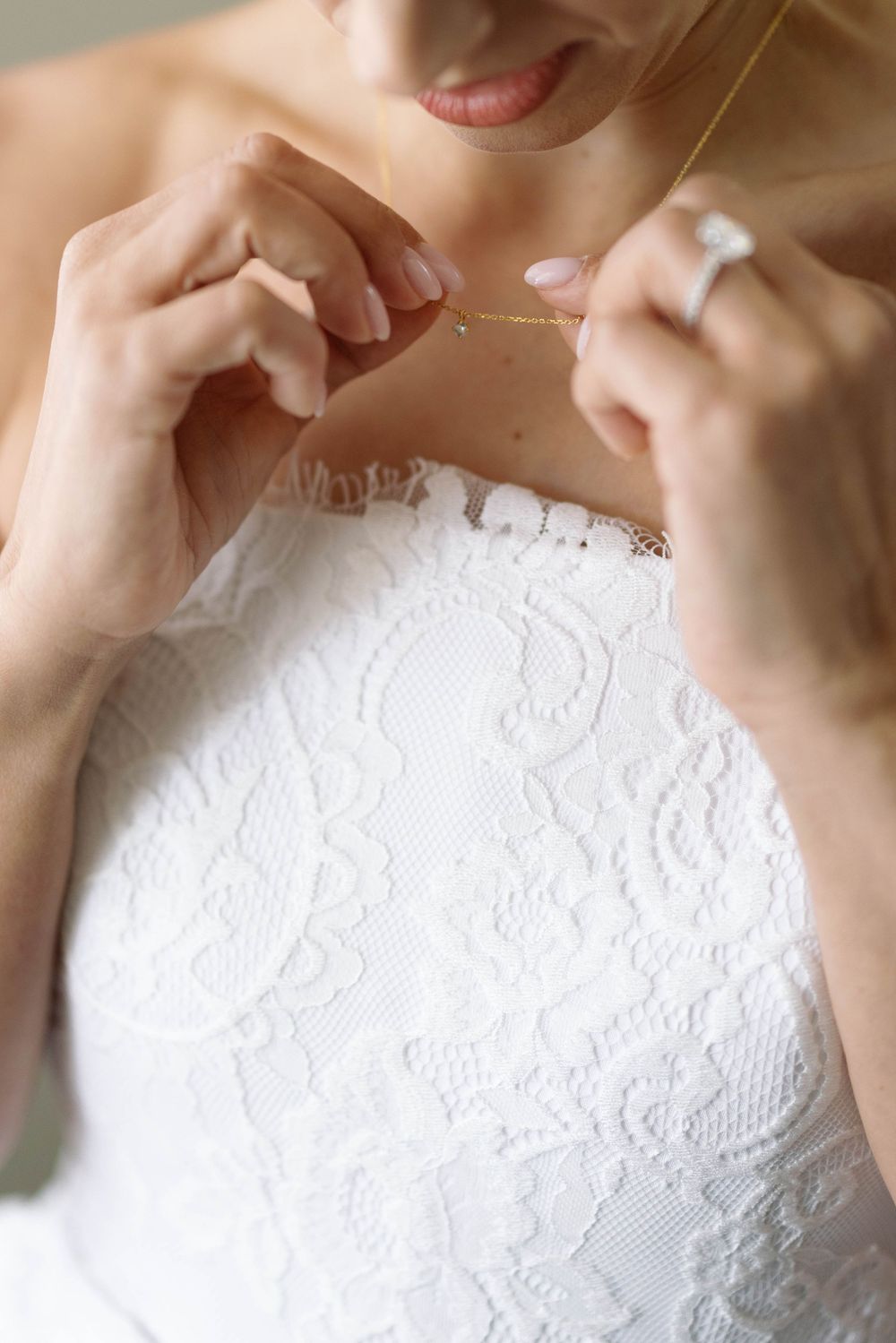 Someone fastens a delicate necklace while wearing an elegant white lace wedding dress.