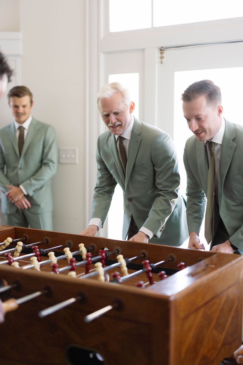 Groomsmen in matching sage green suits gather around a foosball table during wedding celebration.