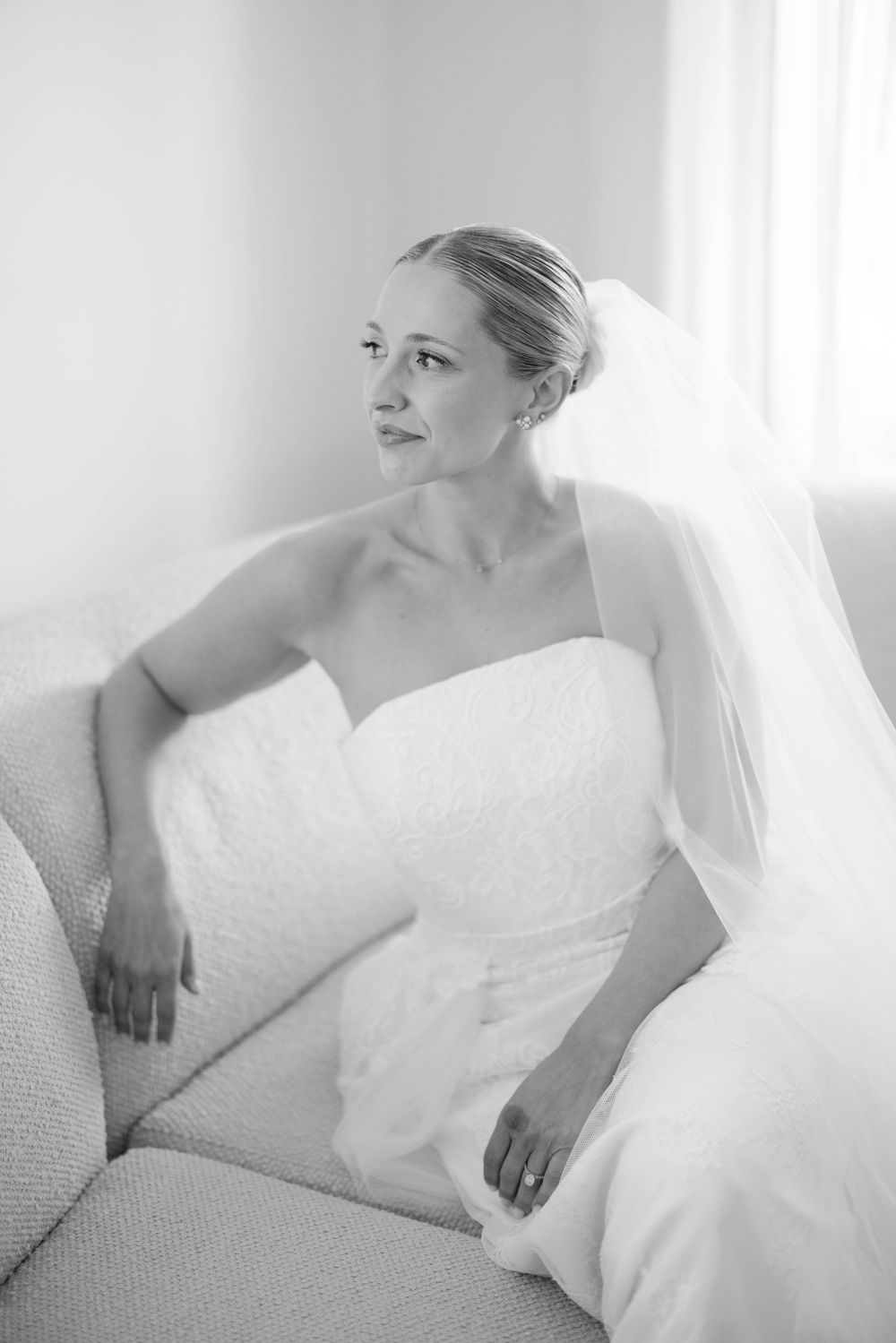A bride in a classic white strapless wedding dress and veil sits elegantly on a light colored couch near a window.