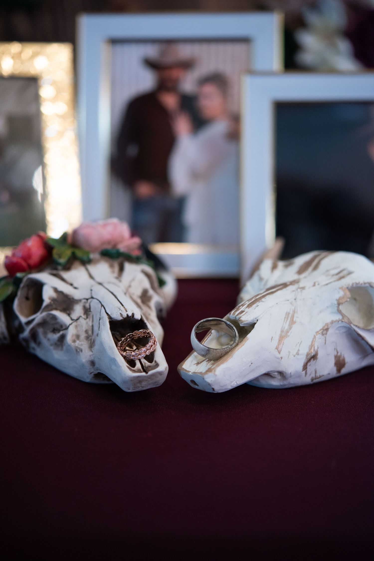 Two white animal skulls displayed with wedding rings and pink flowers against a dark surface with a framed photo in background.