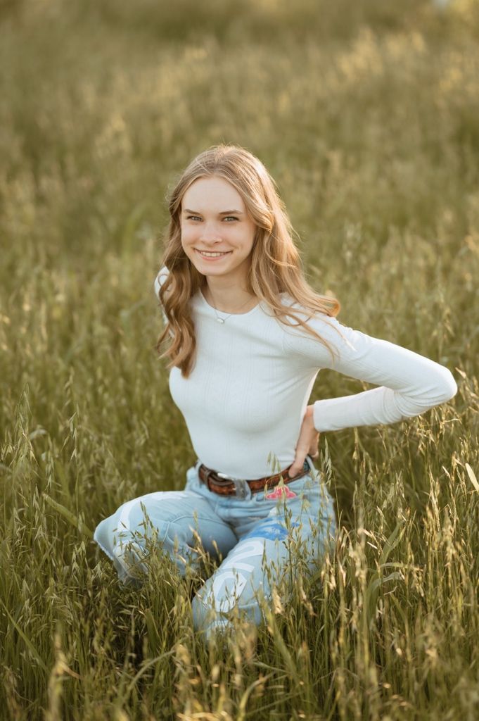 A person in a white long-sleeve top and jeans sits in a sunlit field of tall grass during golden hour.