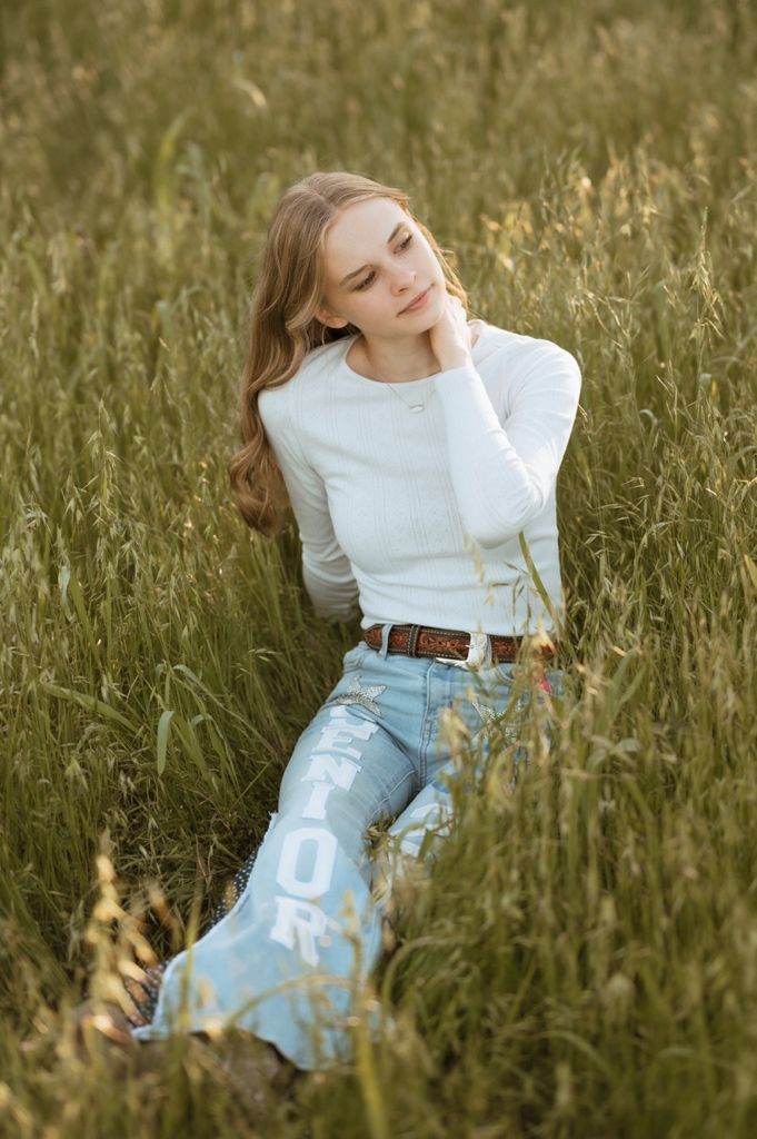 Person in white sweater and distressed jeans sitting in tall grass field during golden hour.