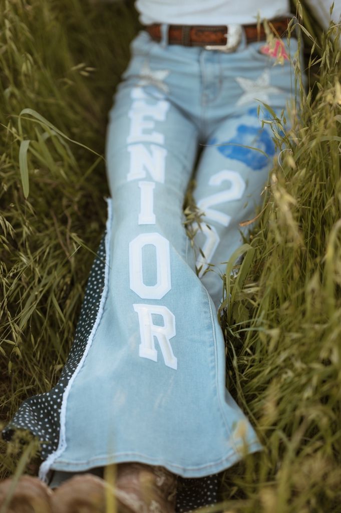 Light wash denim jeans with 'SENIOR' text painted down the leg, photographed in tall grass outdoors.