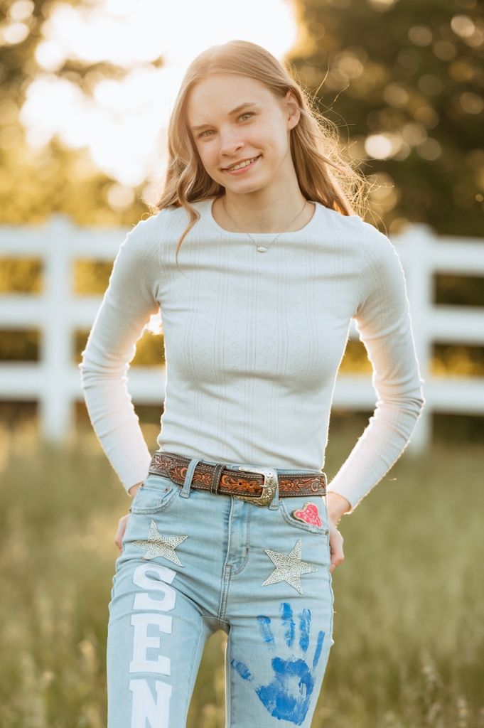 Outdoor portrait showing a white ribbed top and distressed denim jeans with embellishments against a white fence at sunset.