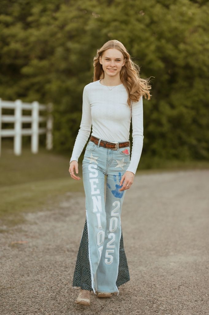 Fashion model wearing white long sleeve top and distressed flared jeans with text print walking on gravel path near white fence.