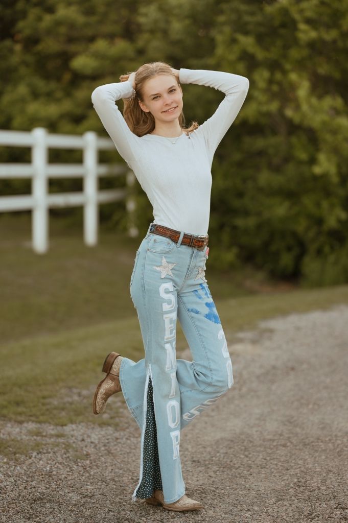Someone in a white sweater and distressed light blue jeans poses playfully on a dirt path with a white fence in background.