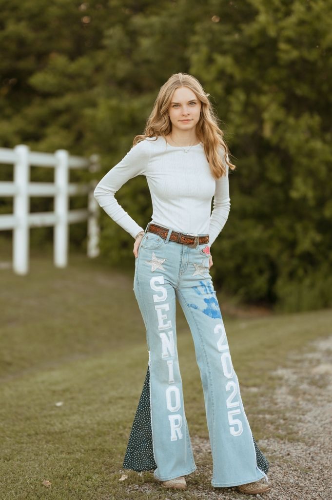Long sleeve white top paired with light wash flared denim jeans with text detailing and brown belt in outdoor setting.