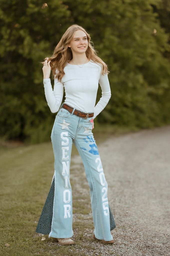 A person in a white long sleeve top and light blue jeans with text lettering stands on a gravel path outdoors.