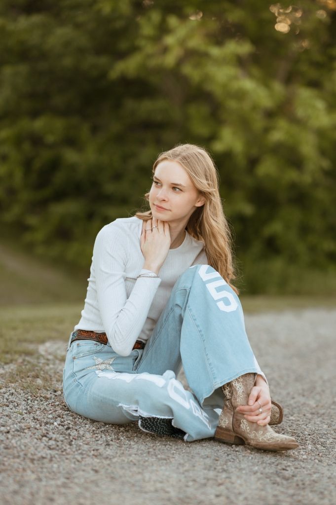 Person in light blue jeans and white sweater sits casually on gravel path with green foliage in background.