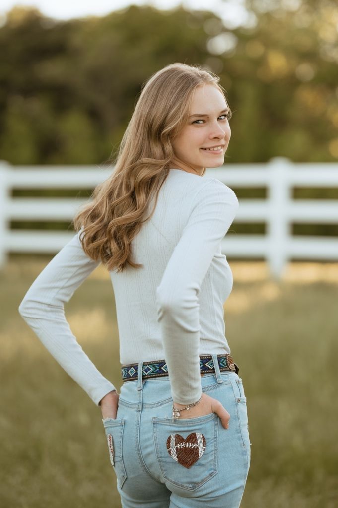 Person in casual attire poses next to white fence at golden hour while holding football and showing playful gestures.