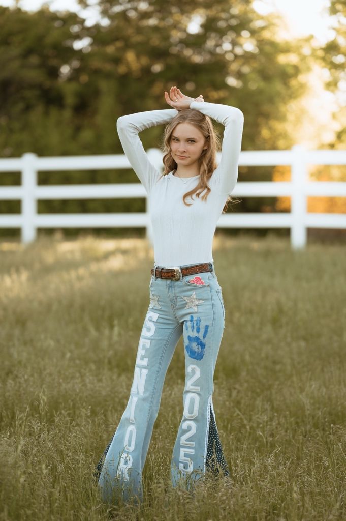 Person in blue jeans and white sweater poses in golden sunset light against white fence and grassy field.
