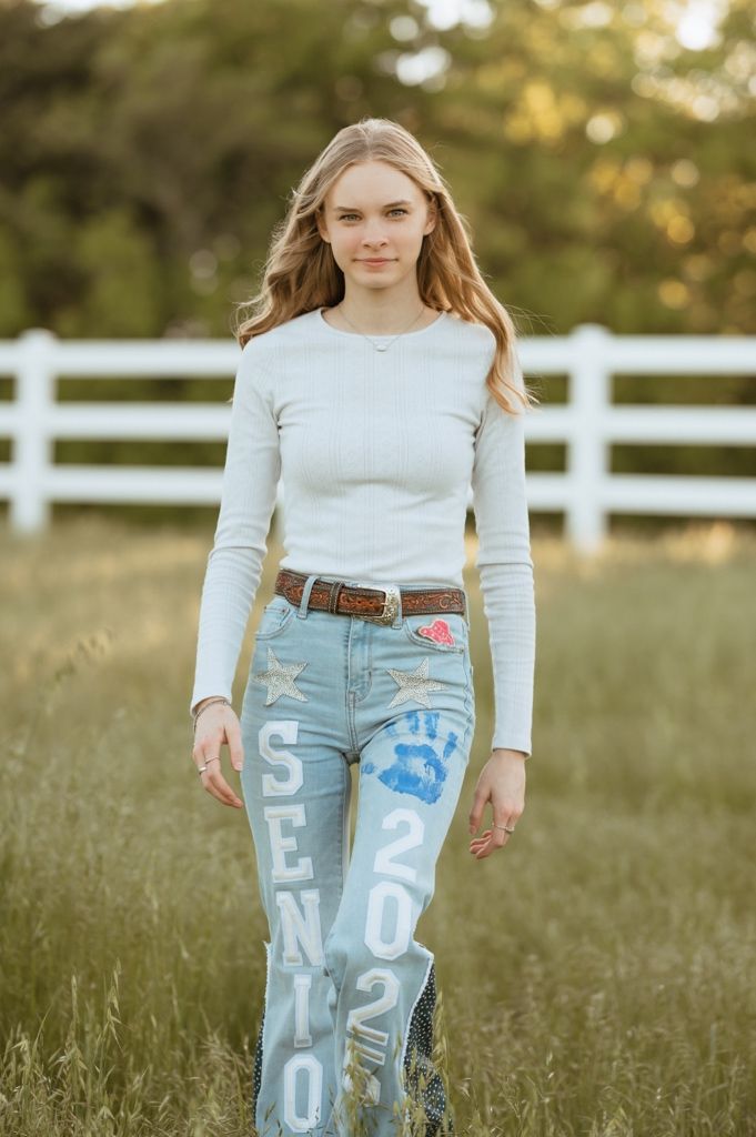 A person wearing a white long sleeve top and painted denim jeans stands in a grassy field with white fencing in background.