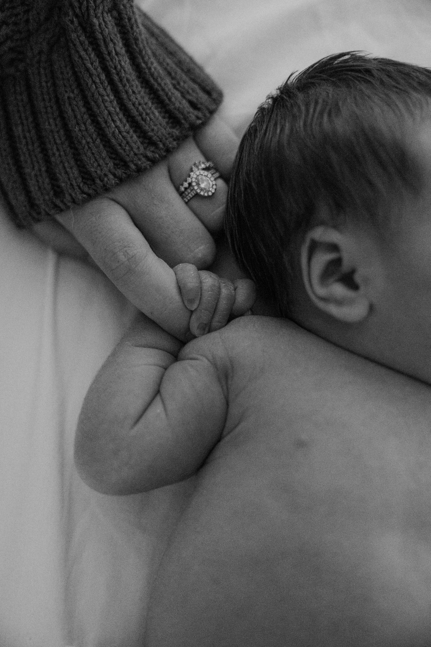 A tender black and white close-up of a baby's hand holding an adult's finger with an engagement ring.