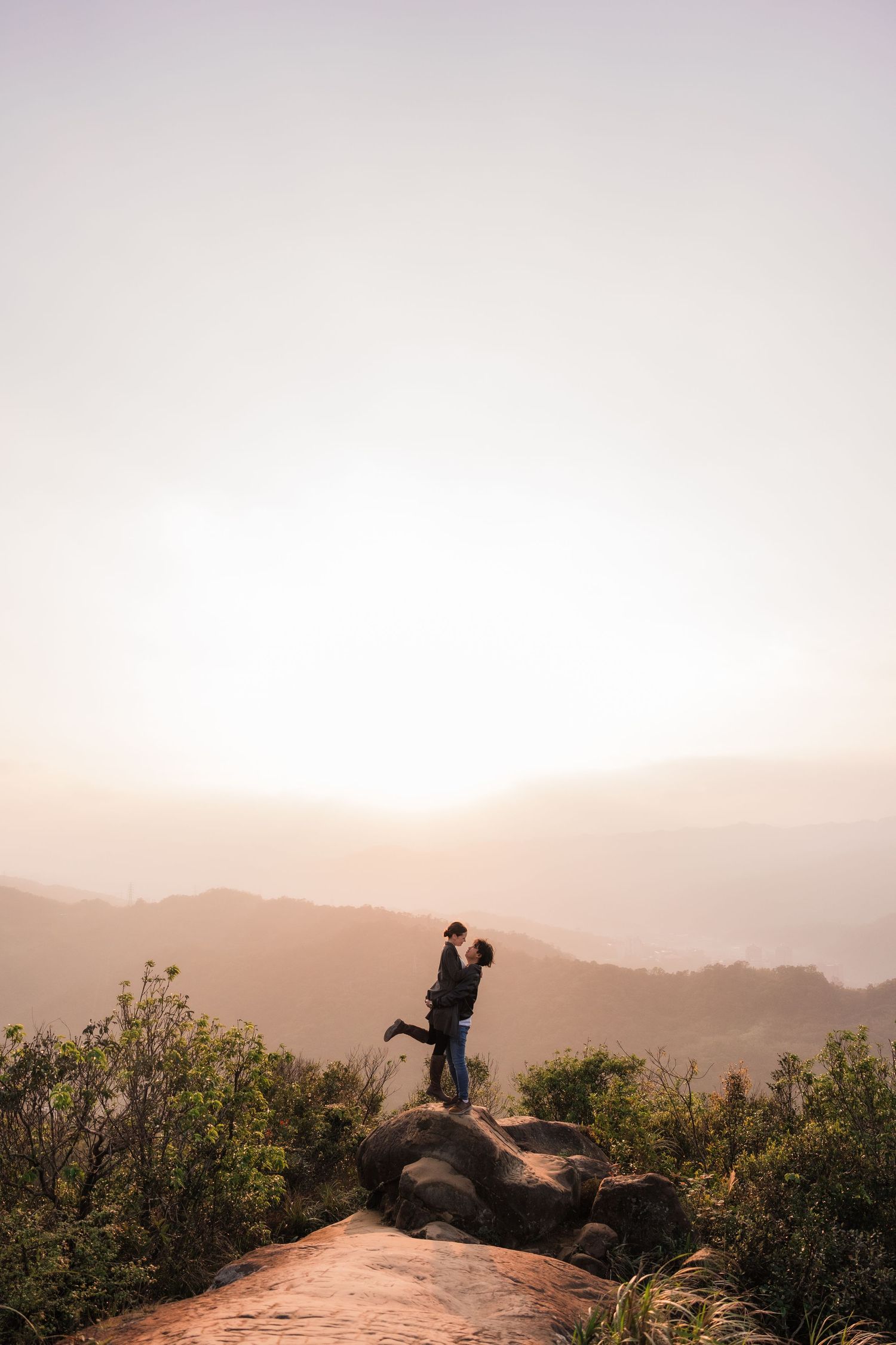 Two silhouettes embrace on a rocky outcrop against a misty mountain landscape at sunset.