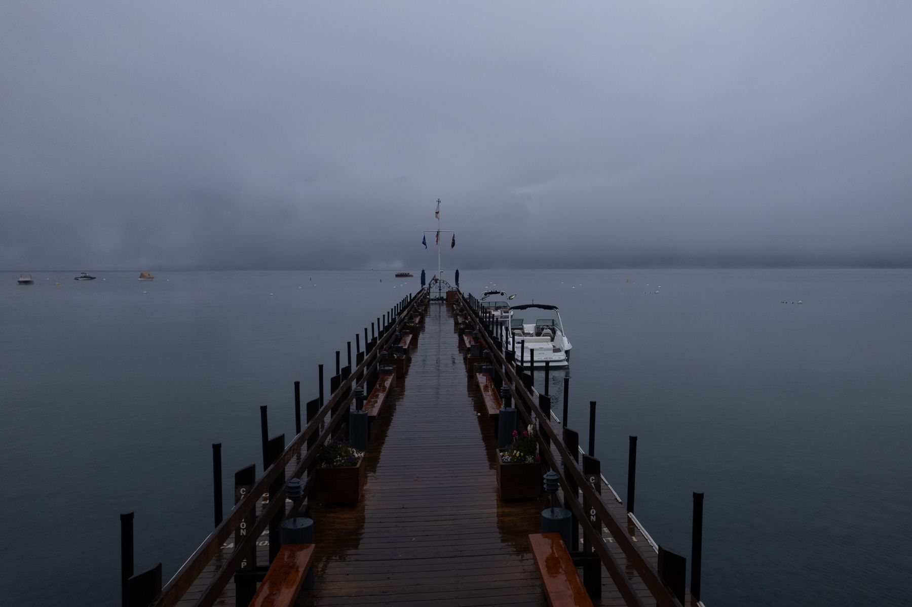 A dark moody wooden pier extends into foggy waters with metal railings under an overcast gray sky.
