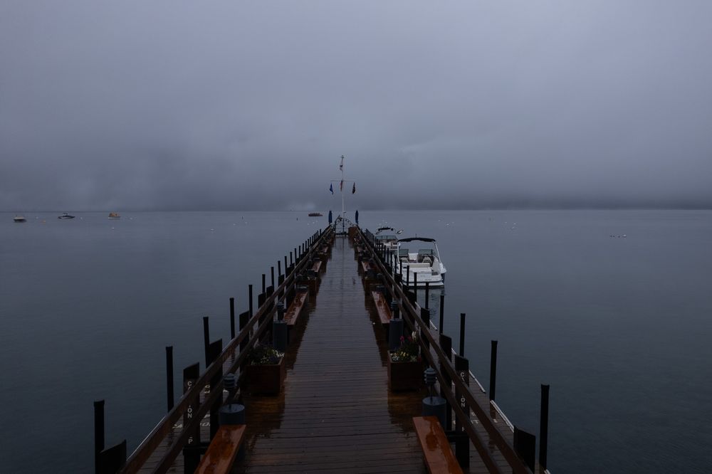 A dark moody wooden pier extends into foggy waters with metal railings under an overcast gray sky.