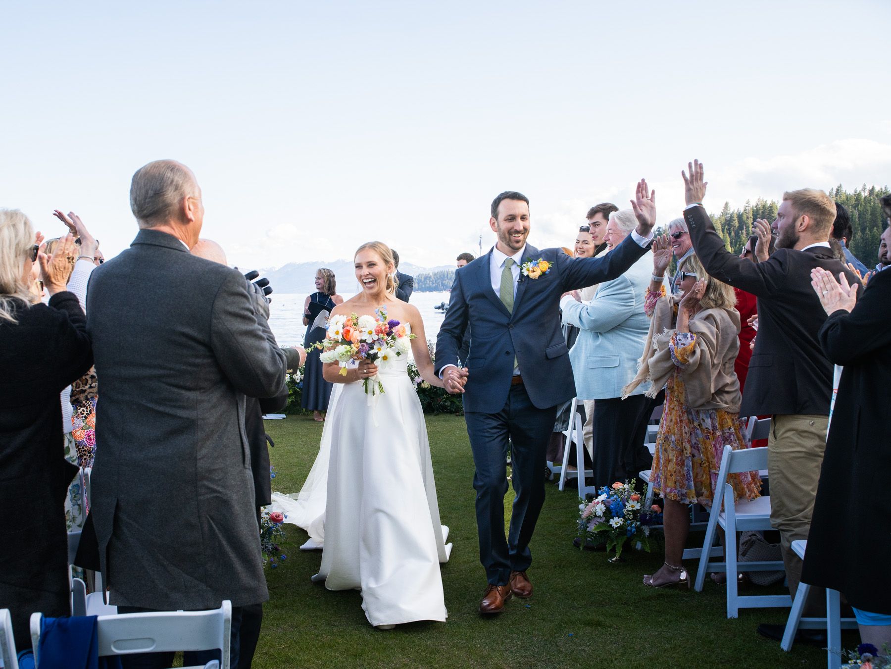 A joyful wedding celebration on a grassy outdoor venue with guests raising their hands as the newlyweds walk down the aisle.