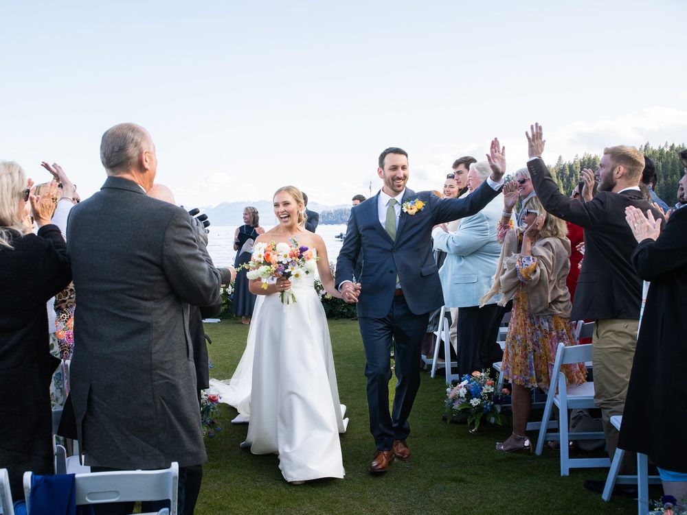A joyful wedding celebration on a grassy outdoor venue with guests raising their hands as the newlyweds walk down the aisle.