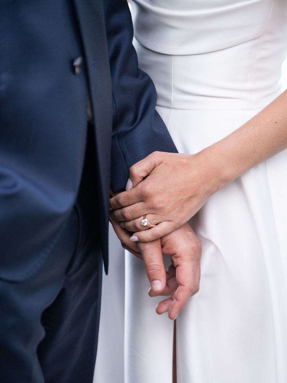 Close-up of newlywed couple's hands with engagement ring, wearing navy suit and white dress.