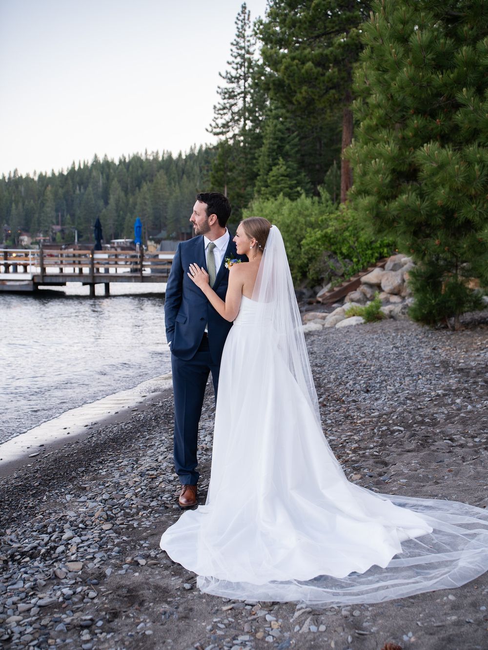 A picturesque lakeside wedding portrait series showing a couple on a rocky beach with tall evergreen trees and a dock.