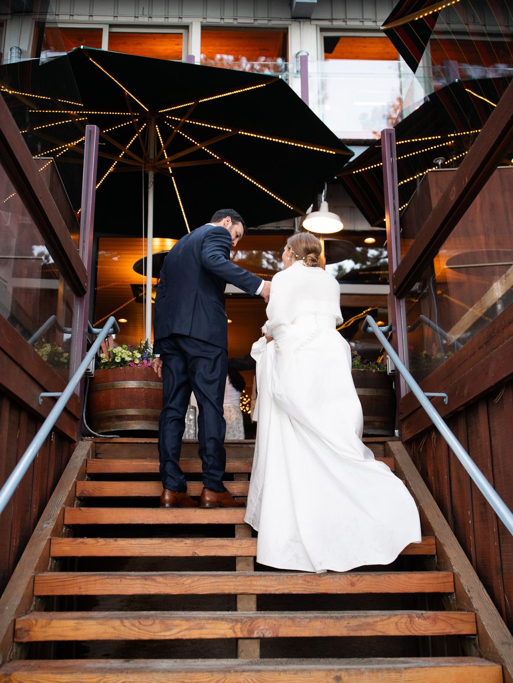 A couple ascends wooden stairs under black umbrellas decorated with string lights at a wedding venue entrance.