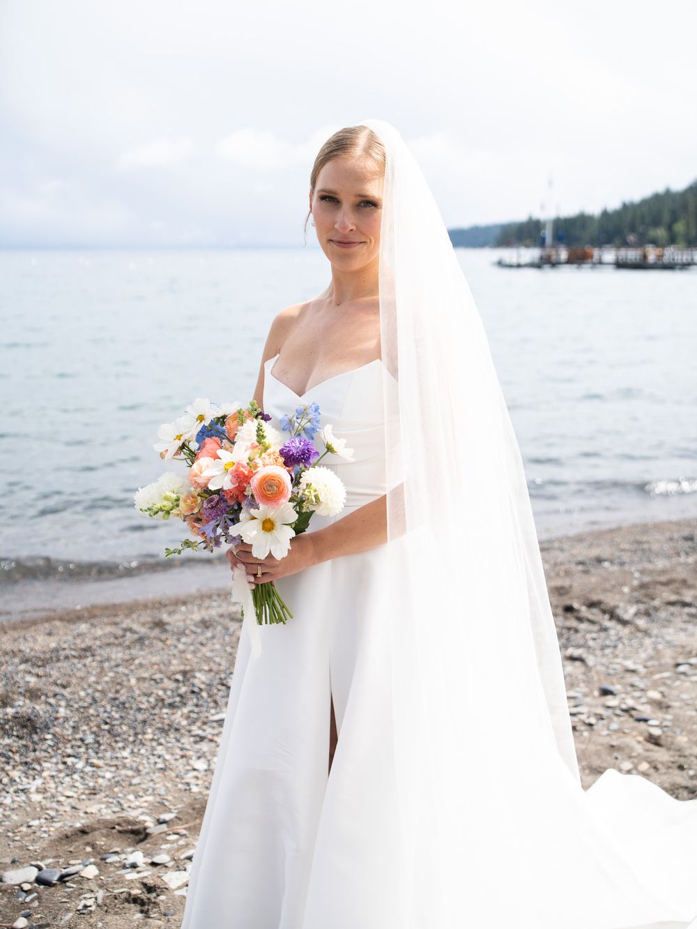 A bride in a flowing white dress and veil holds a colorful bouquet of flowers while standing on a rocky beach.