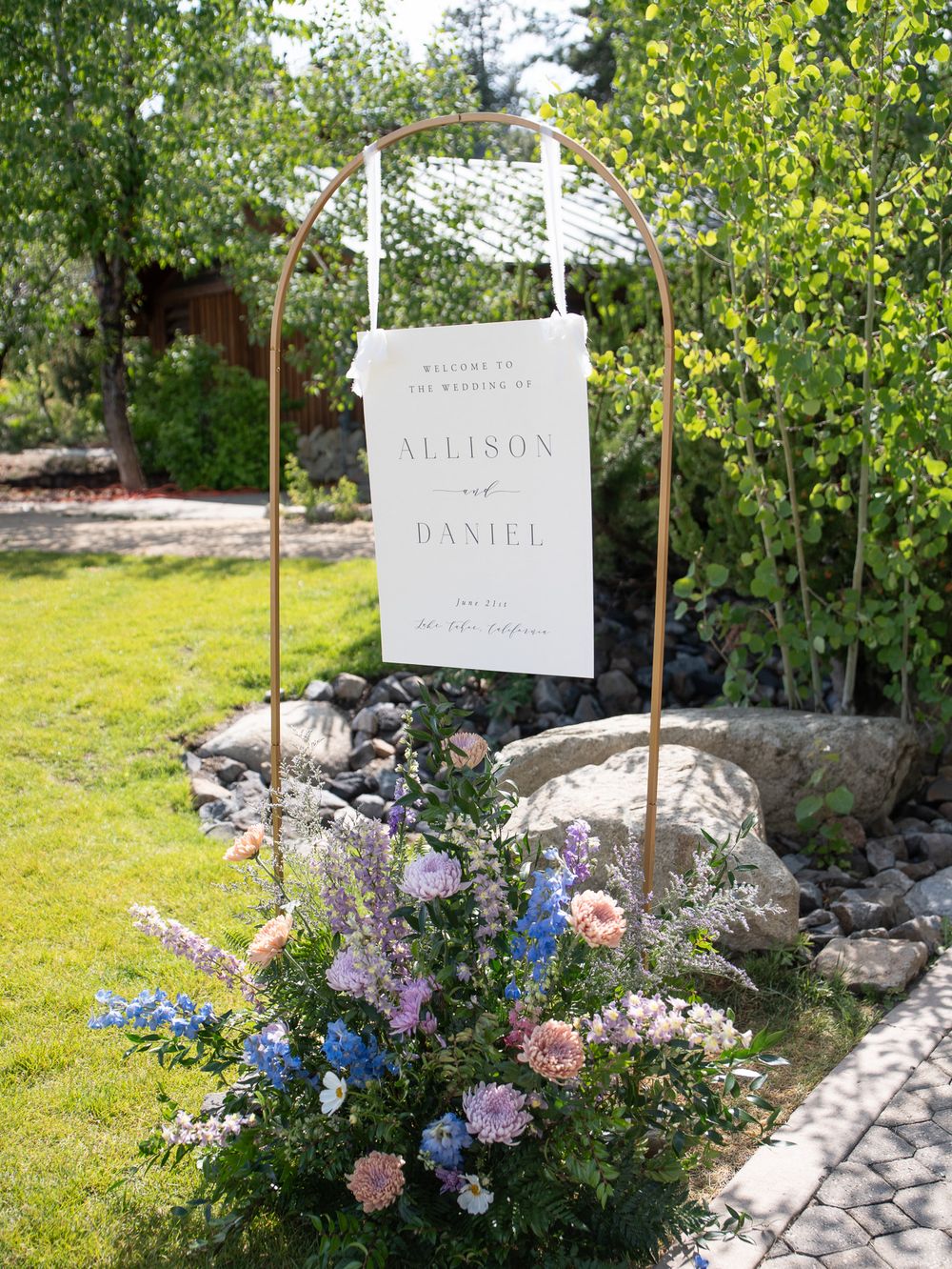 White wedding welcome sign hangs on metal stand with pink and blue flowers blooming at base in garden setting.