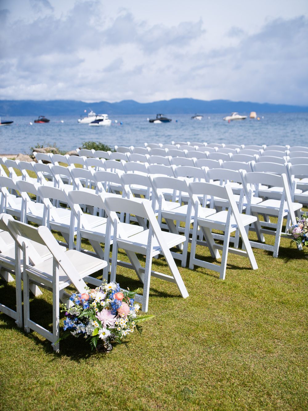 Rows of white folding chairs arranged on grass with mountains and water views provide seating for an elegant outdoor ceremony.