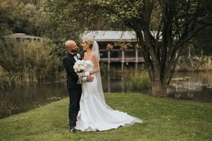 Two people in wedding attire embrace on a grassy lawn beside a pond with a gazebo in the background at Grand Lodge Oaklands.