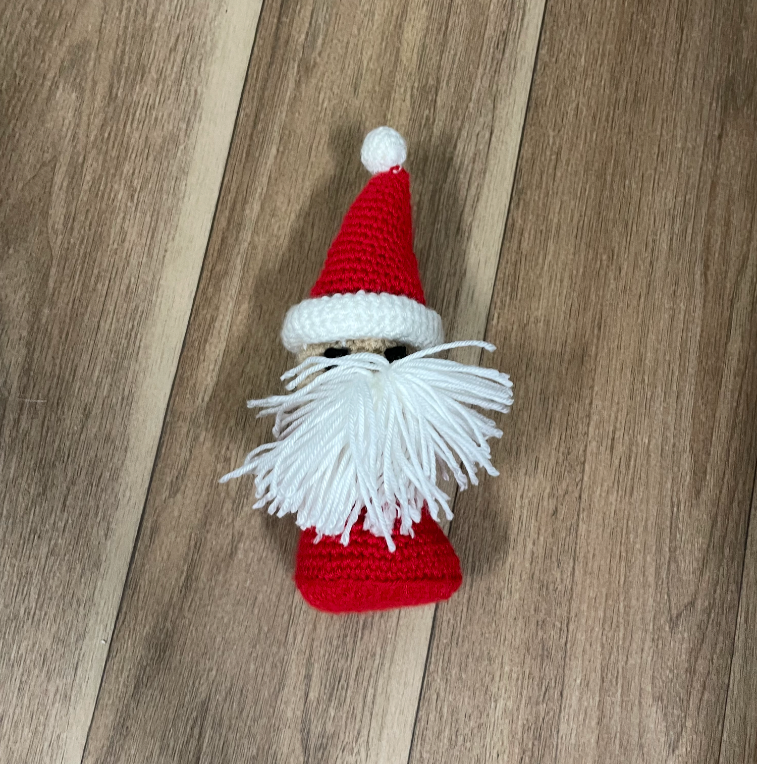 A crocheted Christmas gnome with white beard and red hat on wooden flooring.