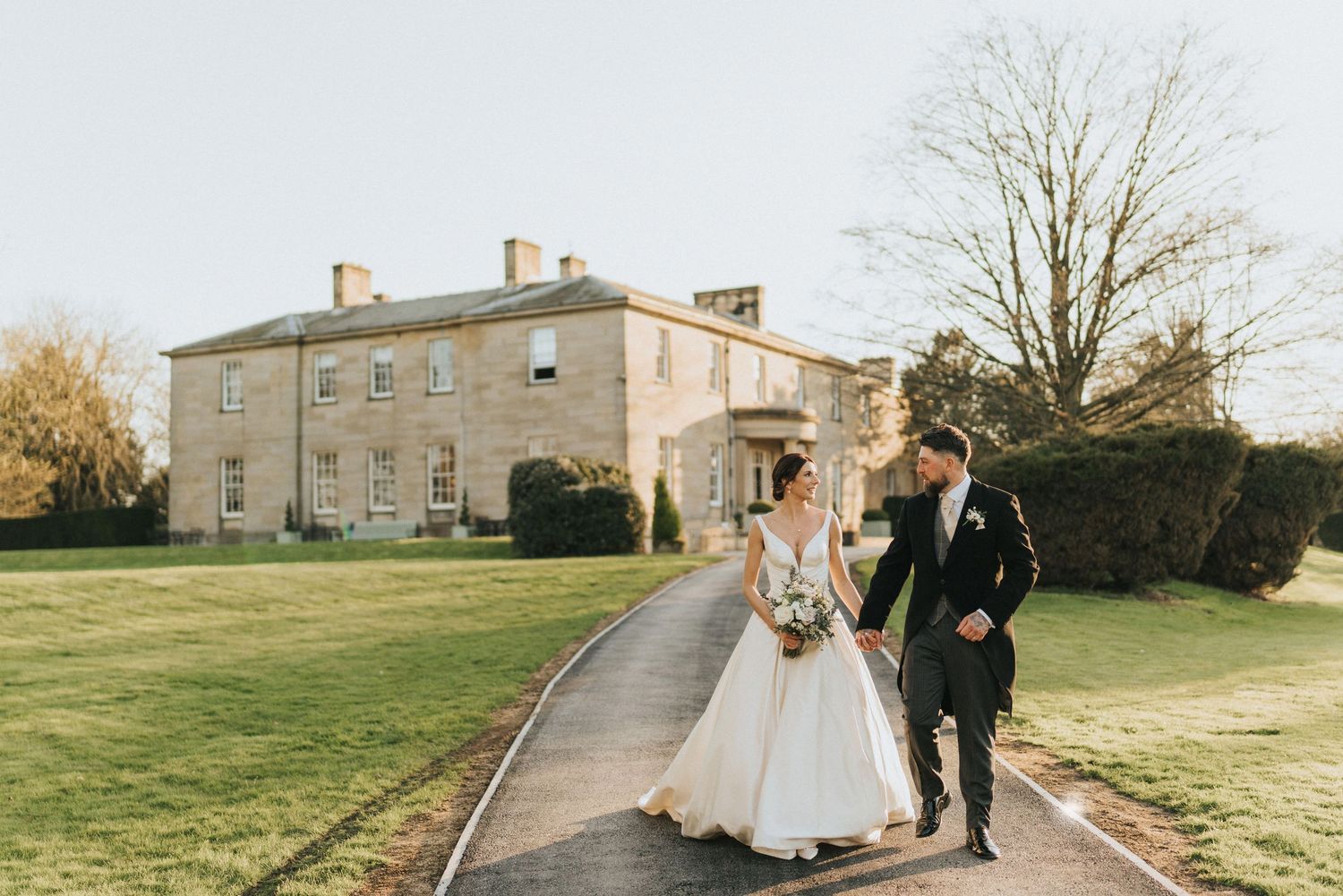 A couple in wedding attire walks along a path in front of Saltmarshe Hall on a sunny autumn day. Image taken by Grimsby photographers.
