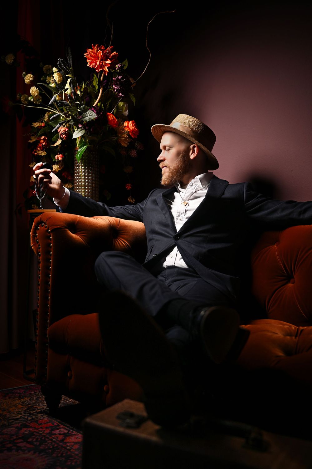 A dark, moody photo series showing a person in a hat reclining on a red velvet couch with flowers in the scene.