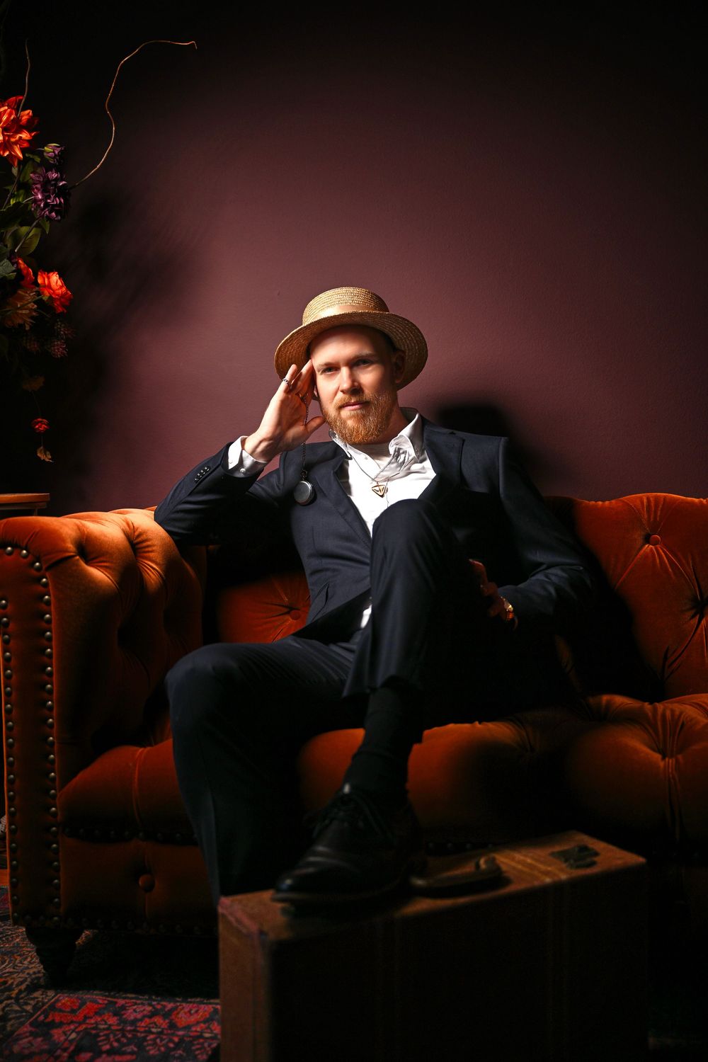 A person in vintage attire sits thoughtfully on a red tufted leather sofa in a moody, dark studio setting.