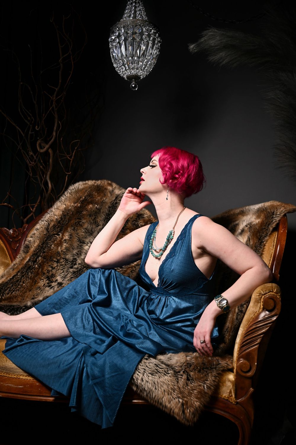 Figure in navy blue dress with bright pink hair reclines on vintage couch under crystal chandelier in dark moody setting.