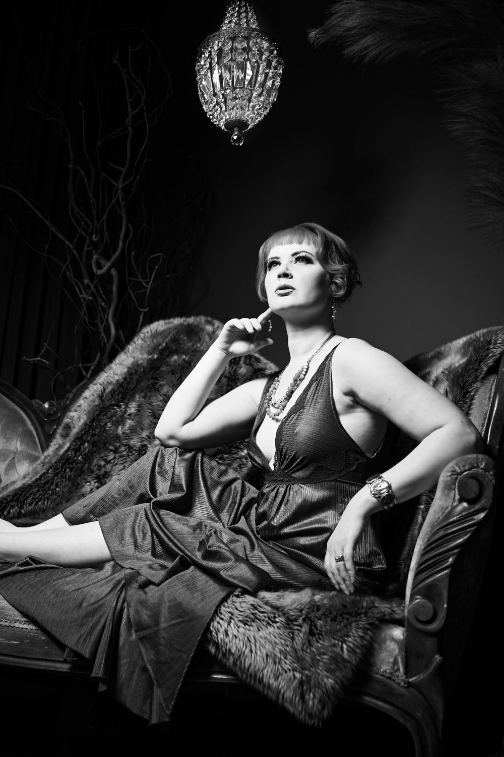 Dramatic black and white photograph showing an elegant figure reclining on a fur-draped chaise beneath a crystal chandelier.