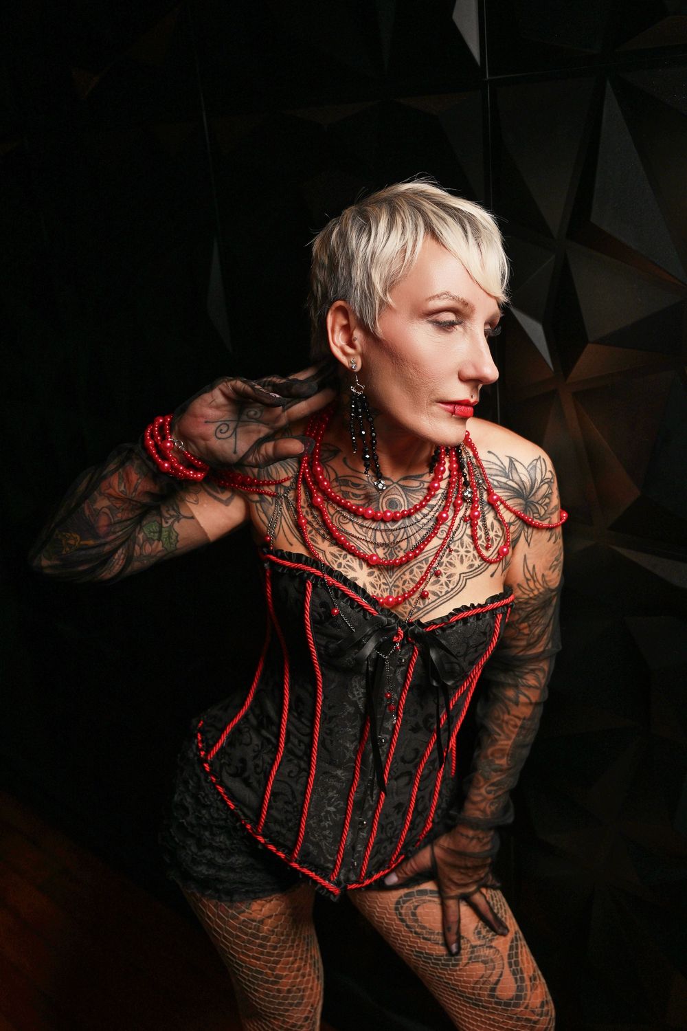 Artistic portrait of someone in a black and red corset with tattoos and red beaded necklaces against dark background.