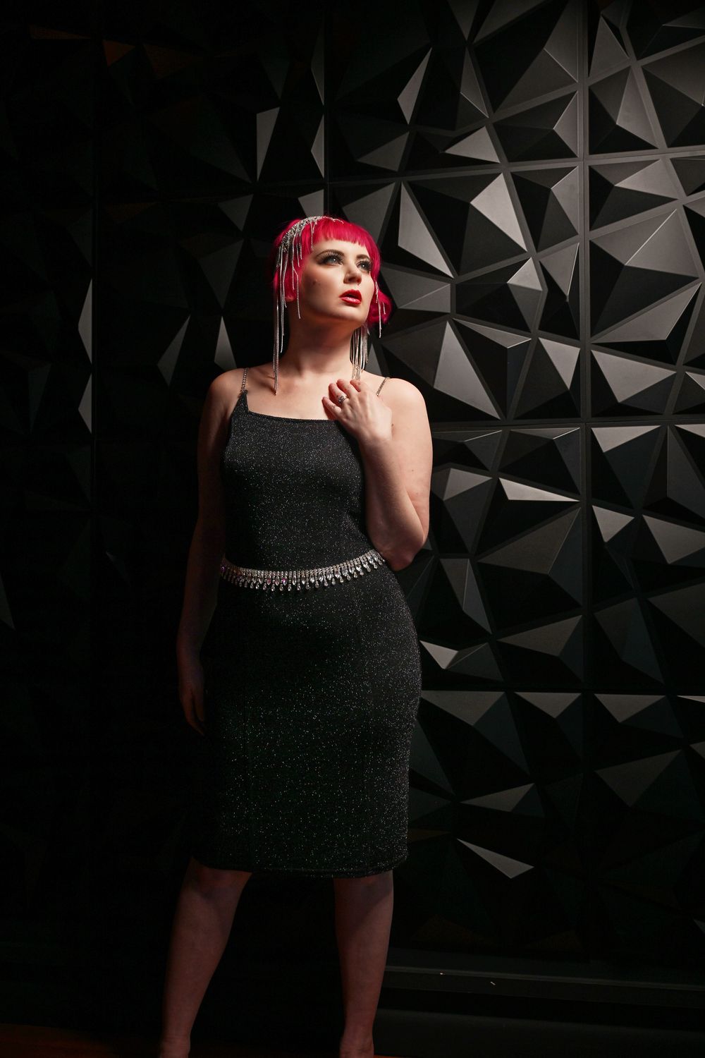 A figure in a black dress with pink hair poses against a dark geometric wall with dramatic lighting.