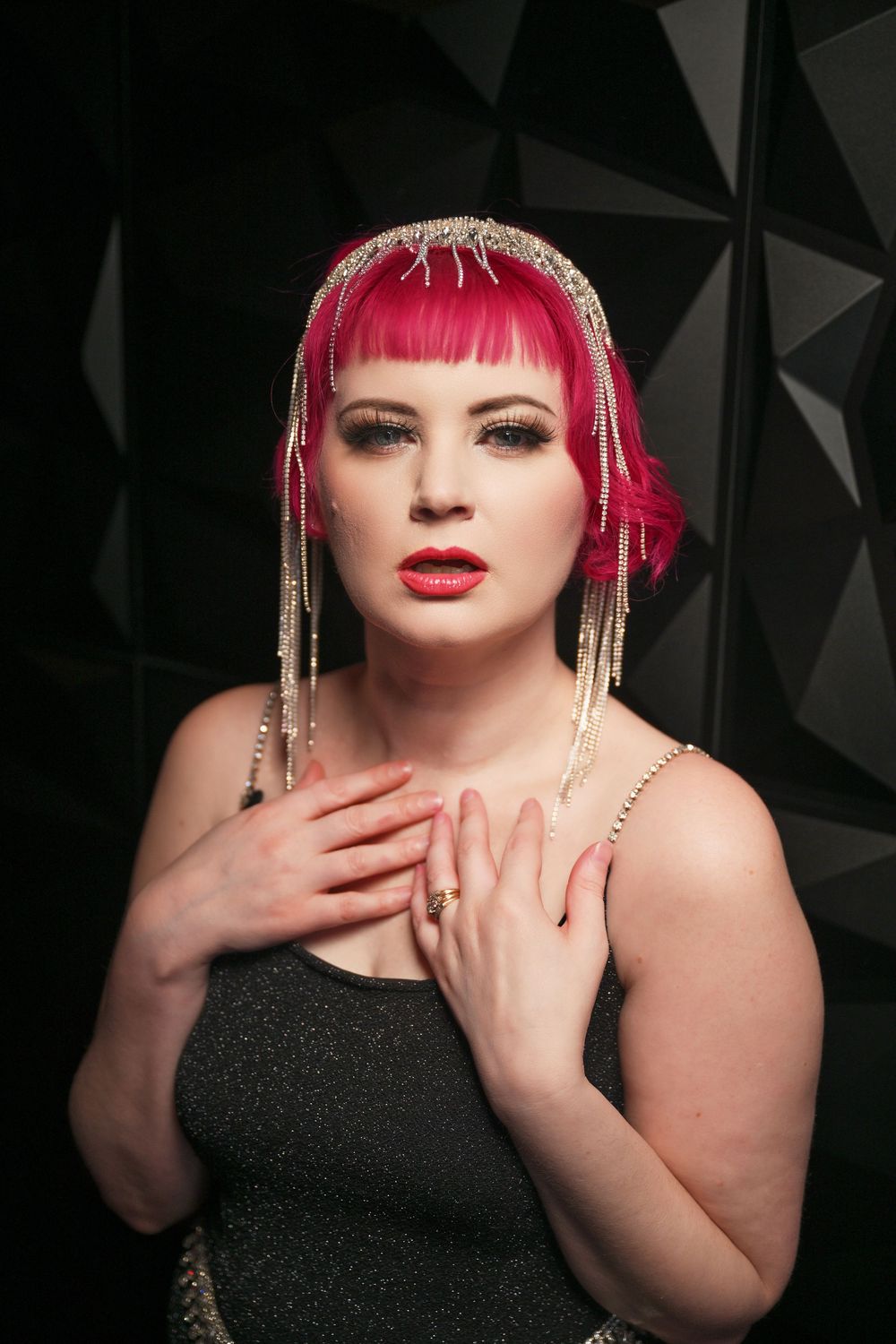 Glamorous portrait with bright pink hair, gold beaded accessories and dark strapless dress against geometric background.
