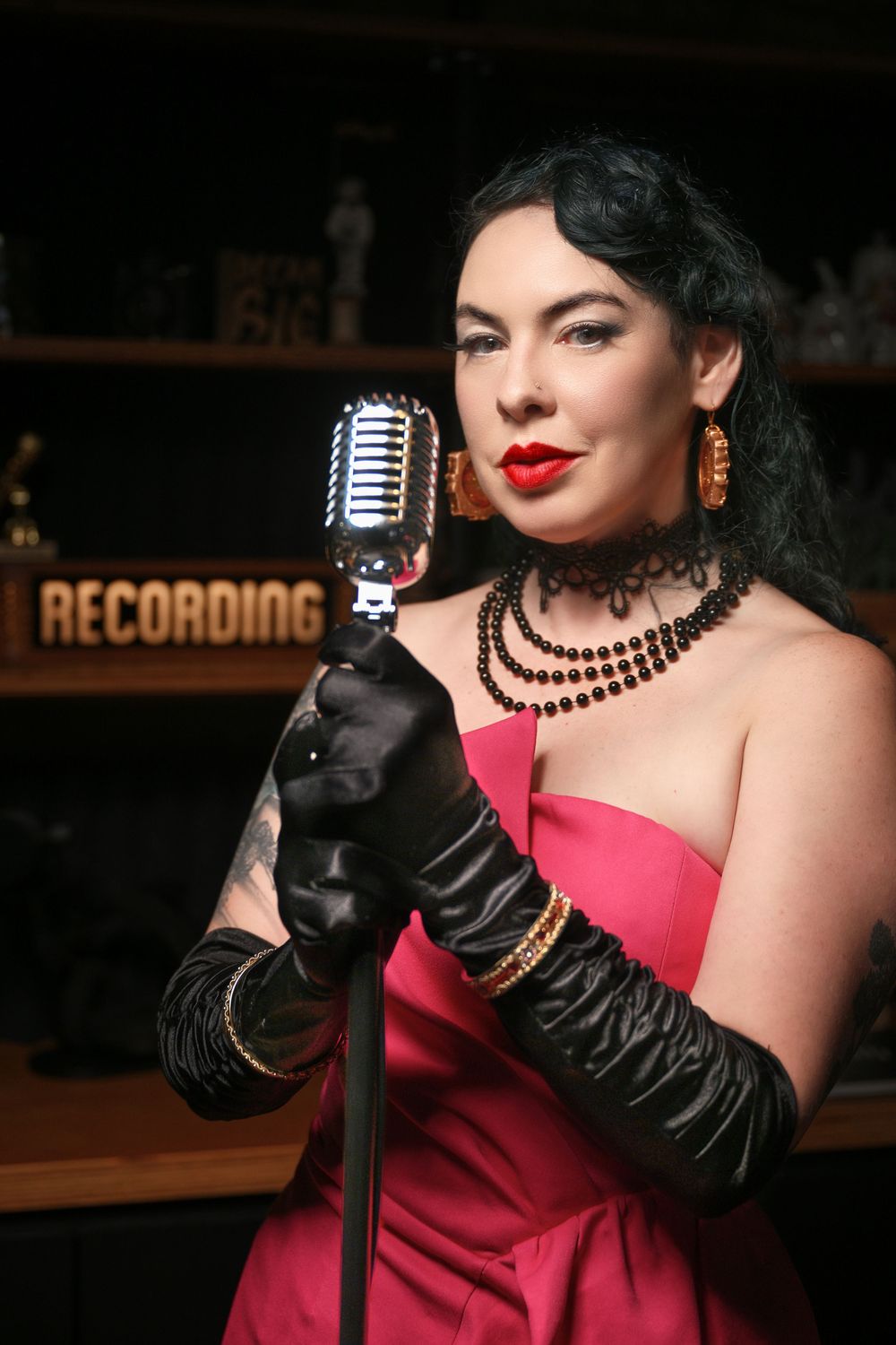 A retro-styled singer in a pink dress and long black gloves performs with a vintage microphone at a recording studio.