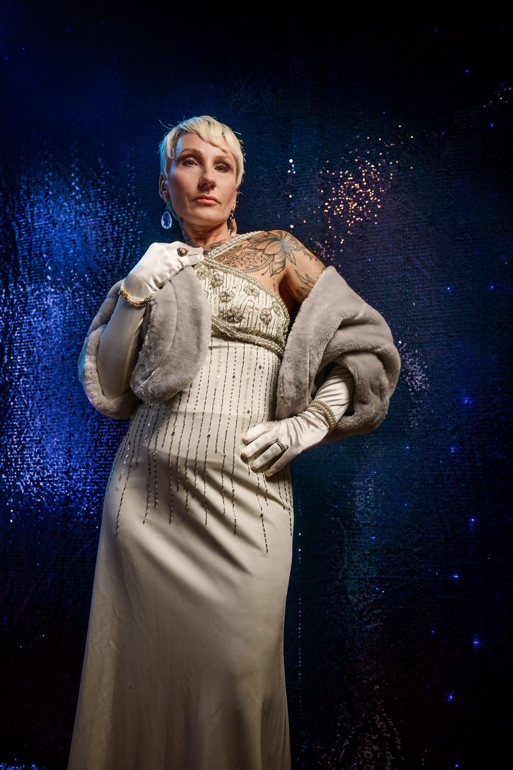 Model poses in elegant cream-colored evening gown with silver embellishments against dark blue sparkly background.