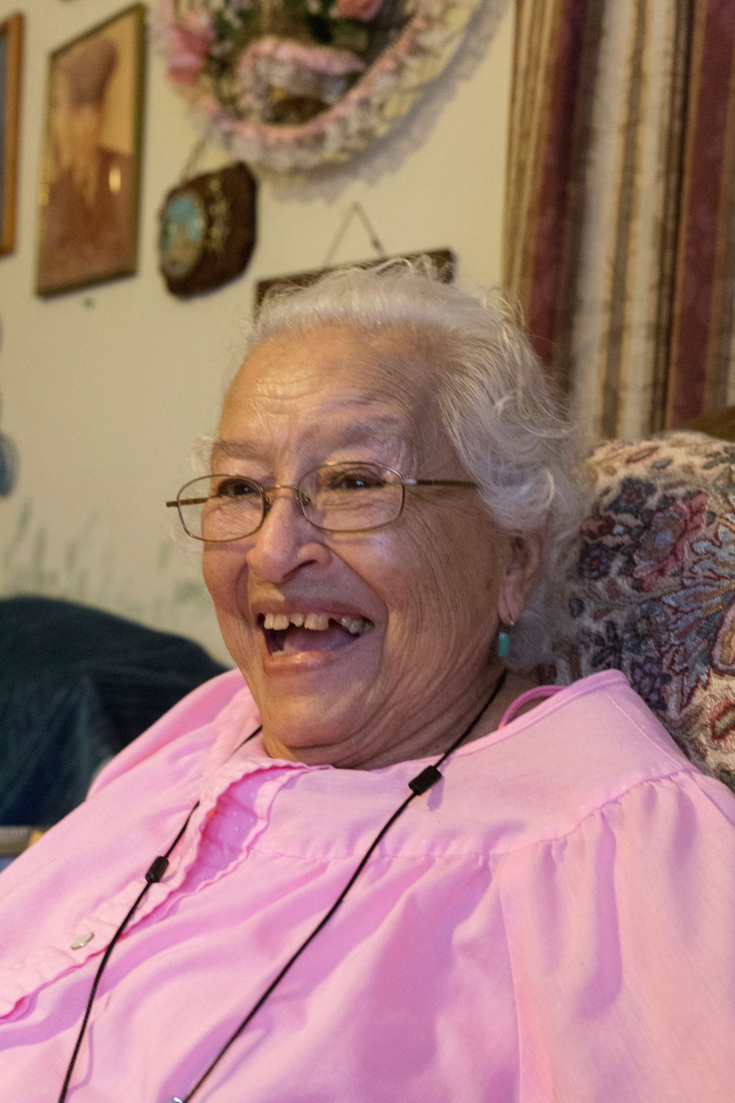 Happy elderly person in a pink shirt and glasses smiling warmly while sitting in a floral chair at home.