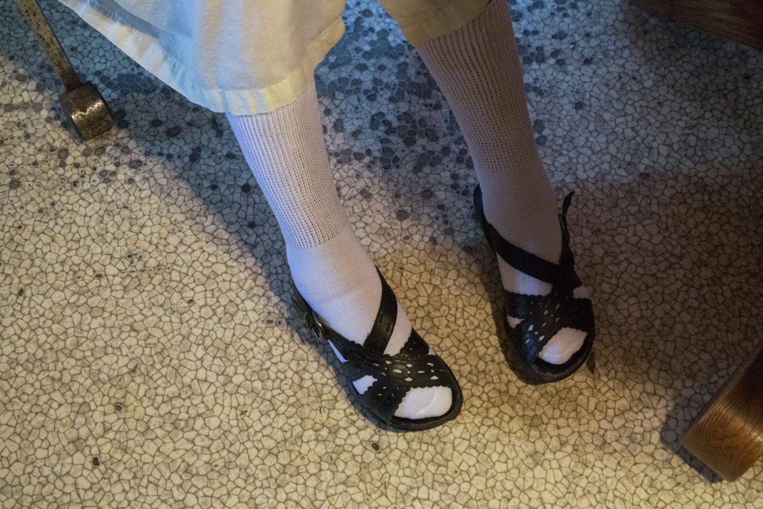 A person wearing white socks and black sandals stands on a speckled carpet floor.