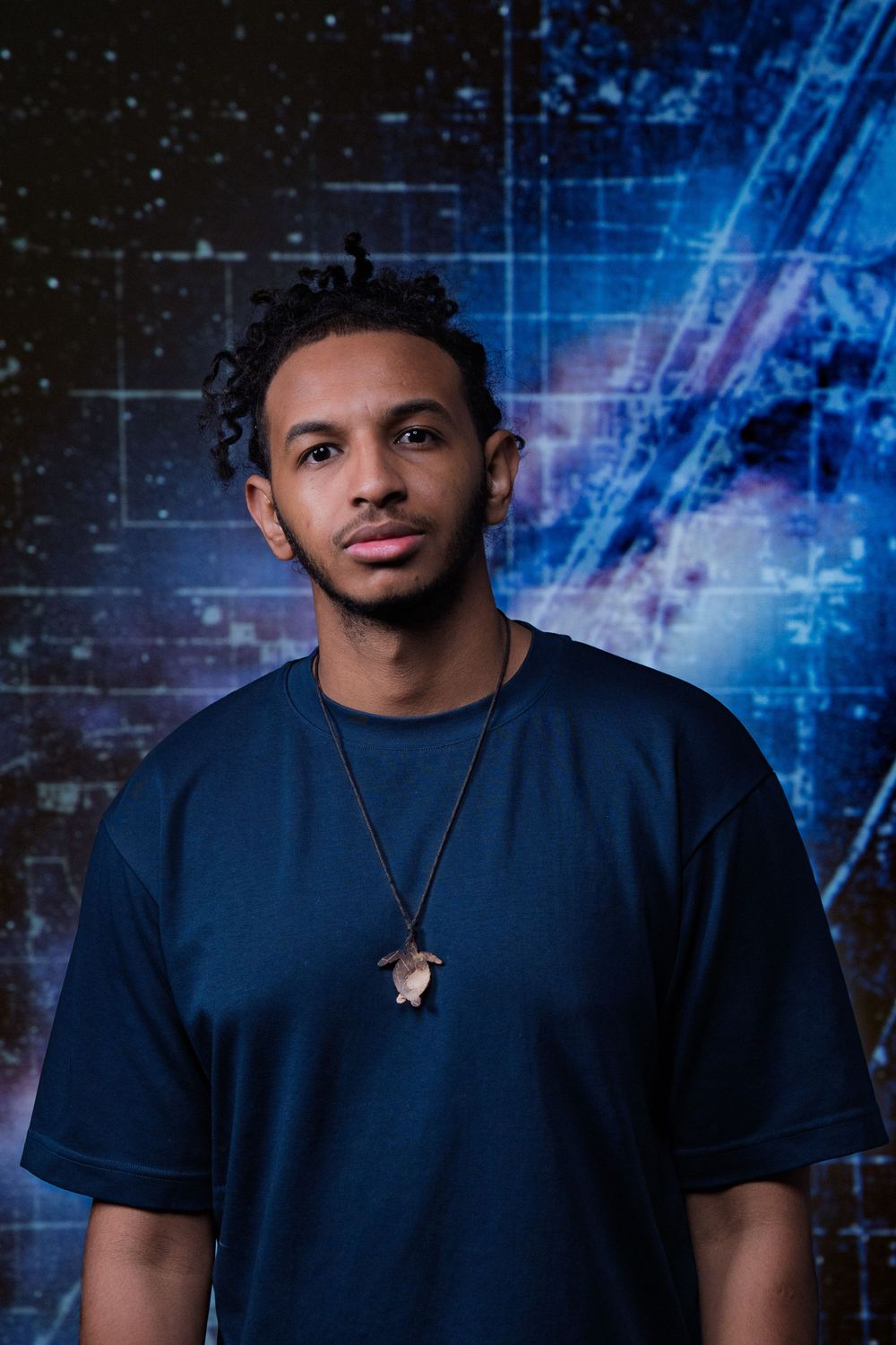 Portrait of a person in a navy blue t-shirt and necklace against a futuristic blue digital background.