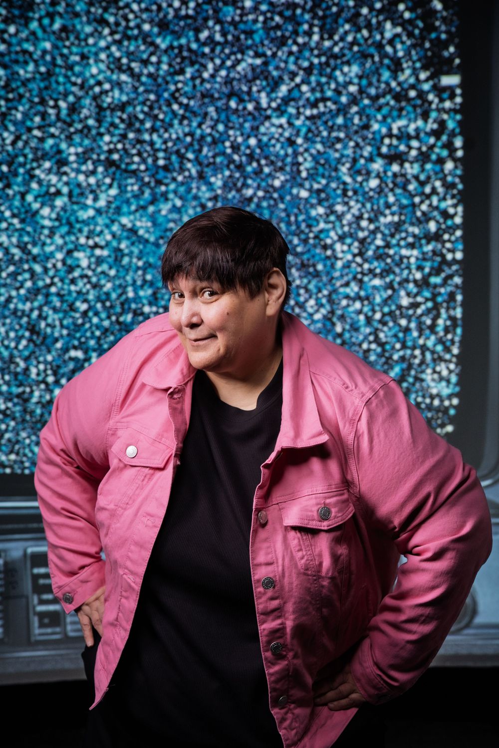 Person in pink jacket poses against sparkly blue background while smiling at camera.