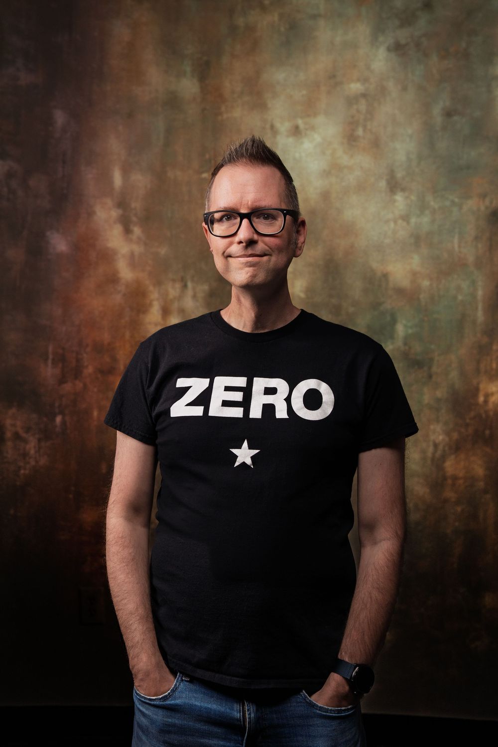 Studio portrait of a person in glasses wearing a black t-shirt with ZERO text and a star against a textured background.
