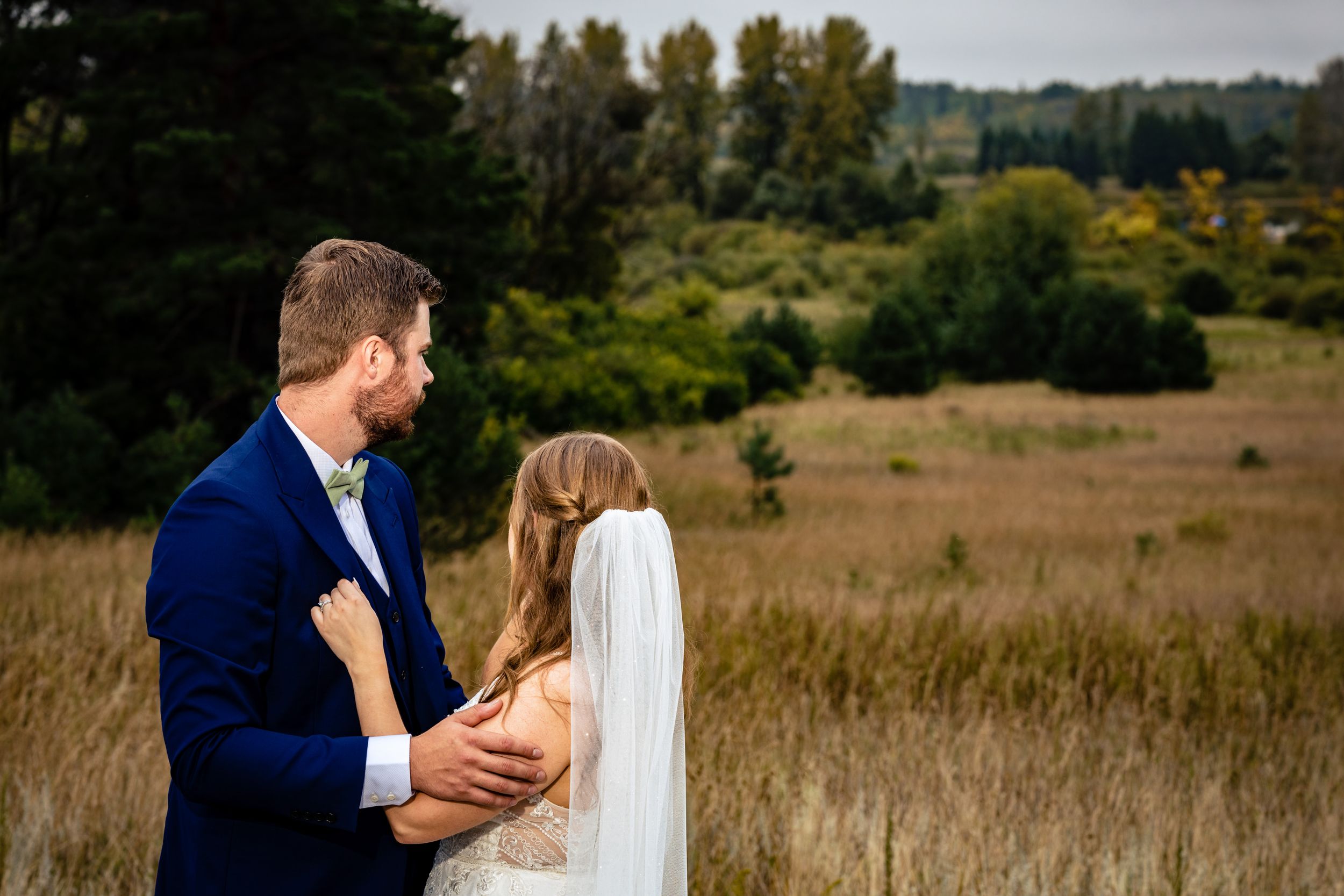 Couple embracing in field with natural landscape and forest in background.