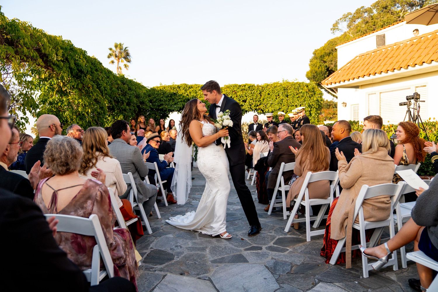 Outdoor wedding guests celebrate with dancing and laughter on a patio overlooking the ocean at sunset.
