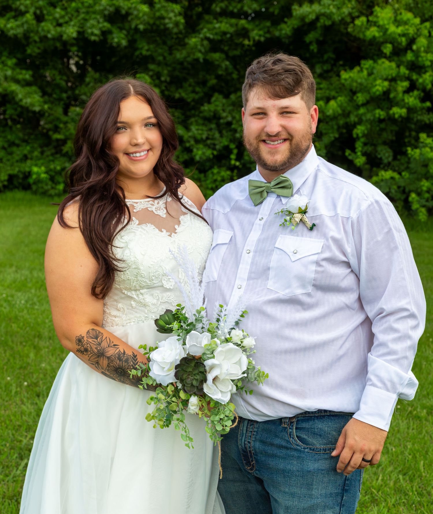 A wedding couple poses together on a lush green lawn with vibrant trees creating a natural backdrop.