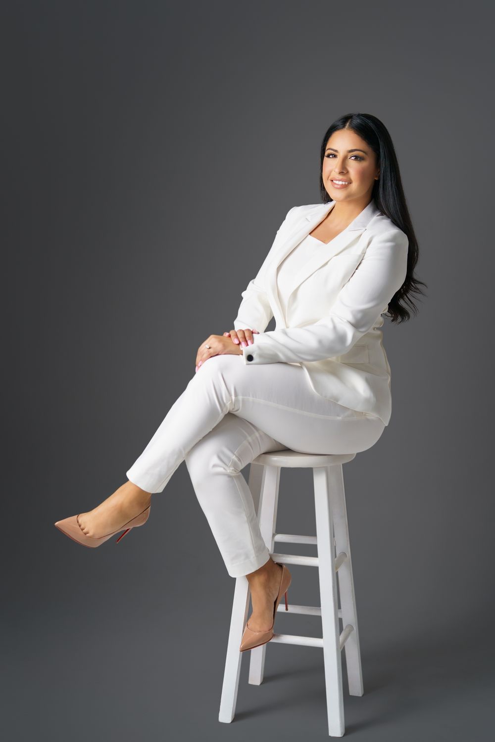 Professional portrait of person in white pantsuit seated on white stool.
