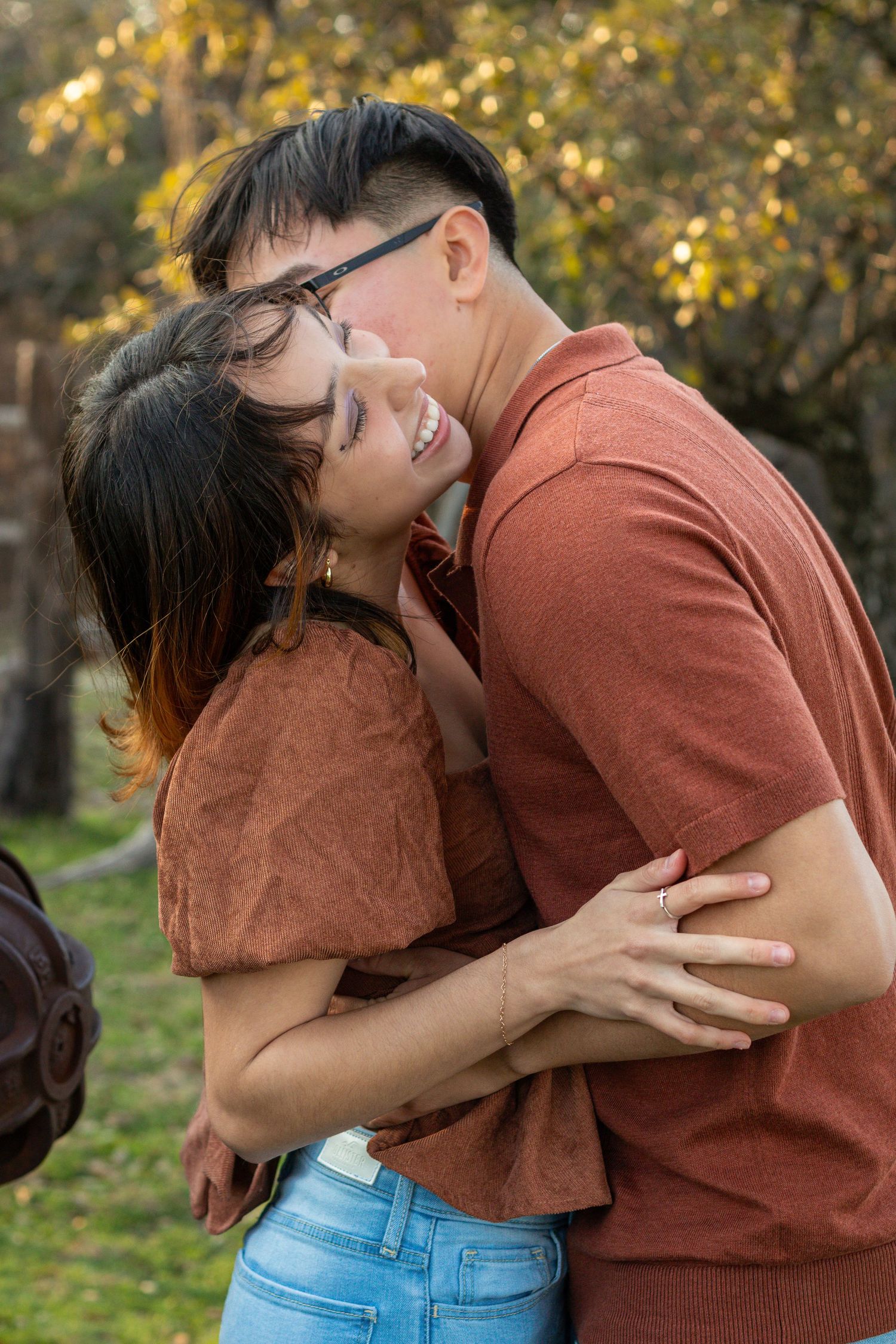 Couple in matching rust-colored tops sharing a tender embrace outdoors with autumn foliage in background.